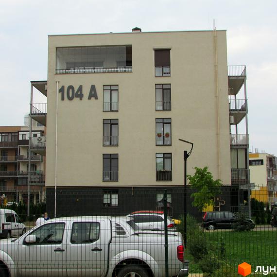 Житловий комплекс Ліпінка: фасад будинку №104 А з вікнами та балконами. Прибудинкова територія з автомобілями та парканом.