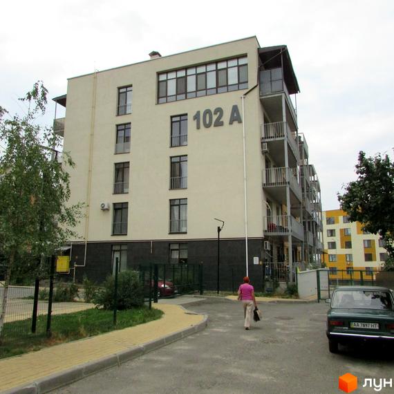 Житловий комплекс Ліпінка: багатоповерховий будинок світлого кольору з балконами та великими вікнами. Поруч дорога, тротуар, зелені насадження та припаркований автомобіль.