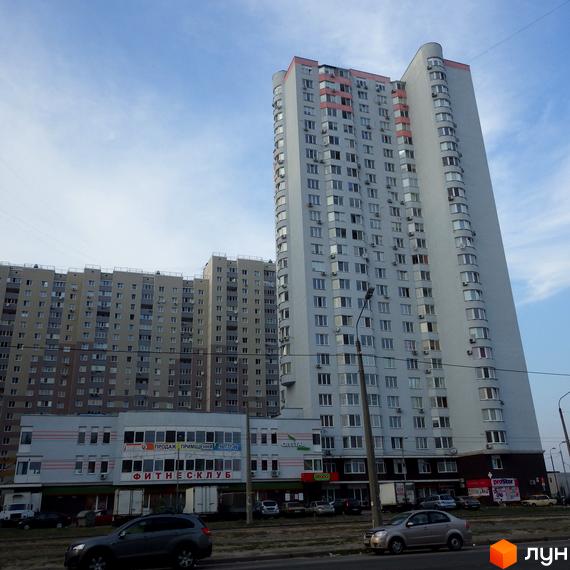 Багатоповерховий житловий будинок світло-сірого кольору з рожевими акцентами. На перших поверхах розташовані комерційні приміщення, поруч дорога.