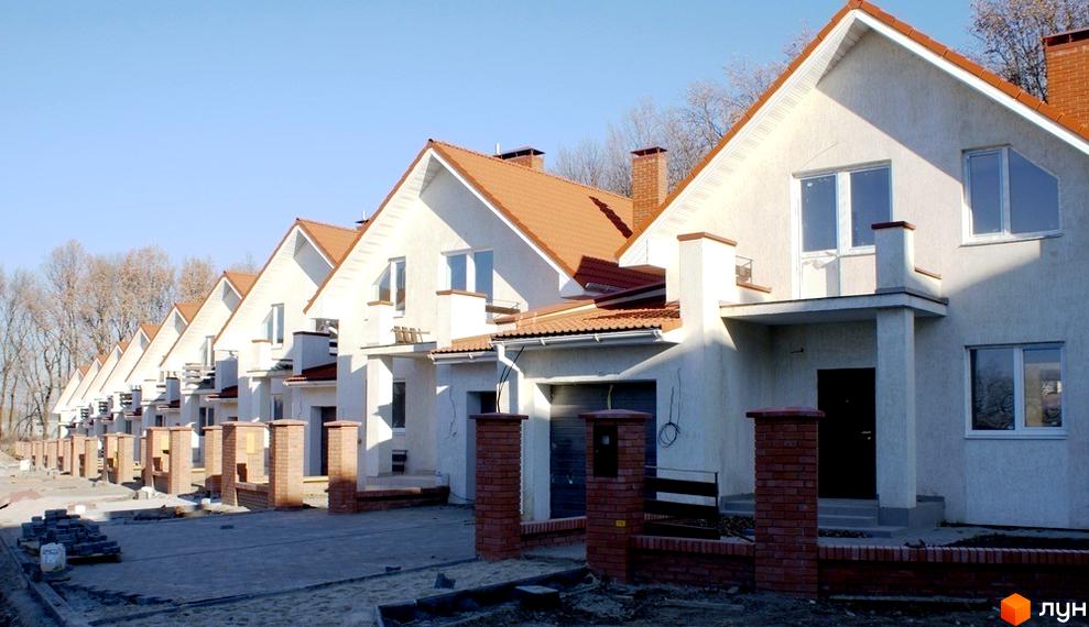 Ряд білих будинків з коричневими дахами. Перед будинками розташовані цегляні стовпчики та доріжки з бруківки, що веде до входу.