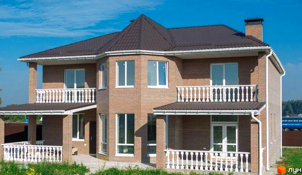 Двоповерховий будинок з балконами та терасою, облицьований цеглою. Навколо будинку зелена трава, а на задньому плані блакитне небо.