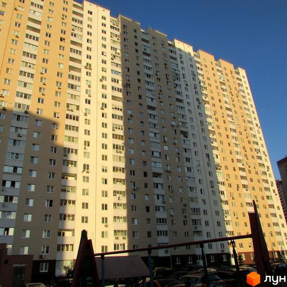 Фасад житлового комплексу на вулиці Урлівській, 38а, з численними вікнами та балконами, що виходить на блакитне небо. Частина будинку освітлена сонцем, а інша в тіні.