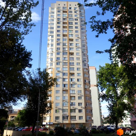 Висока світла новобудова з багатьма вікнами та вертикальними акцентами. Навколо будинку зелені дерева та парковка з автомобілями.