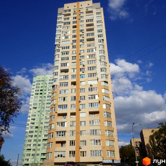 Високий житловий будинок бежевого кольору з помаранчевими смугами та численними вікнами на вул. Феодосійська, 2. На задньому плані видно світло-зелений будинок під блакитним небом.