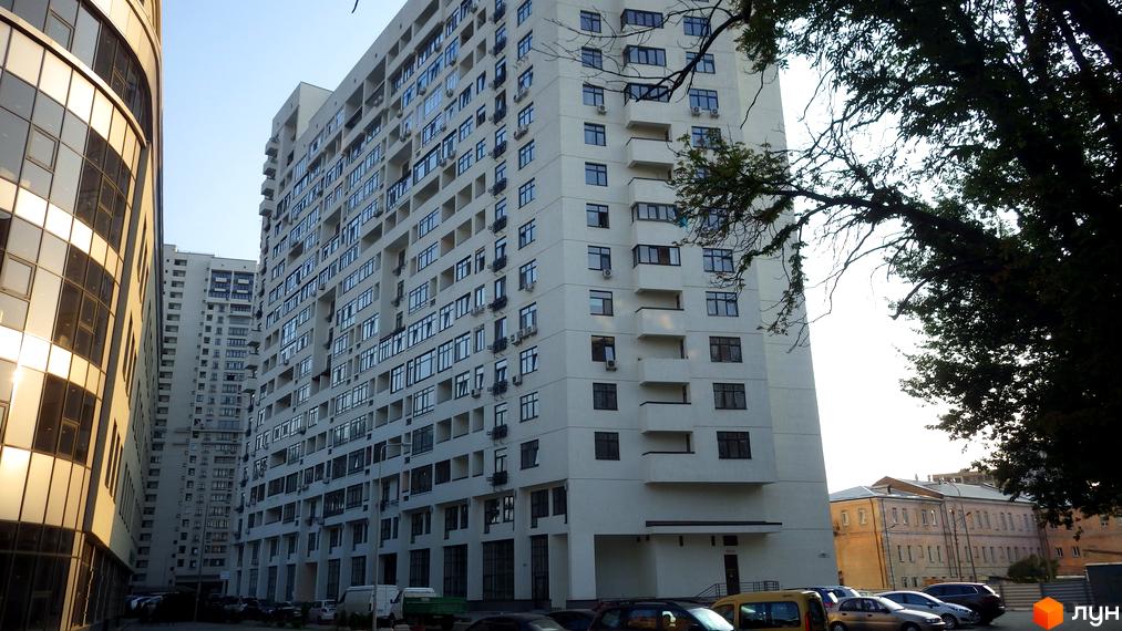 Багатоповерхова світла будівля Житлового комплексу Панорама на Печерську з численними вікнами та балконами. На прибудинковій території припарковані автомобілі та ростуть дерева.