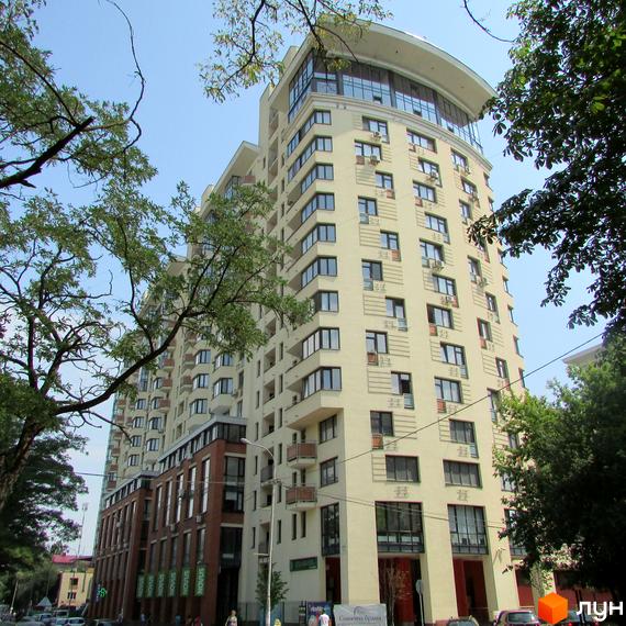 Багатоповерховий будинок Житлового комплексу Сонячна брама зі світлим фасадом, численними вікнами та балконами. Навколо будинку ростуть дерева, видно вулицю з автомобілями та пішоходів.