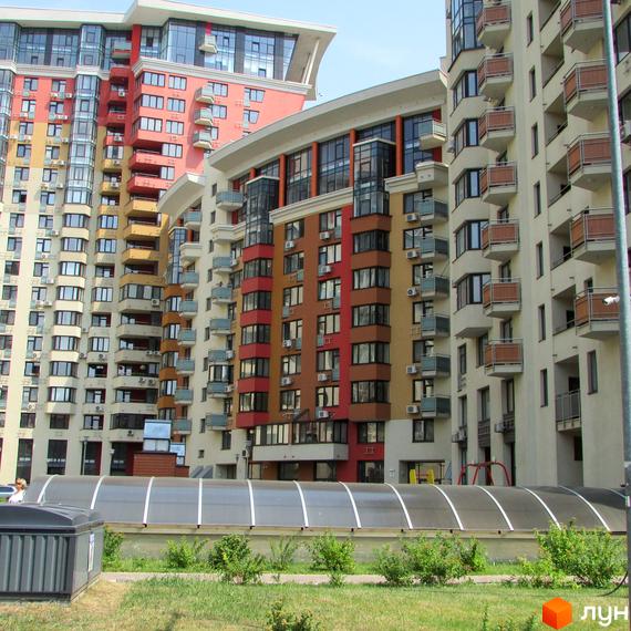 Житловий комплекс Сонячна брама з багатоповерховими будинками бежевого, червоного та коричневого кольорів, балконами та великими вікнами. На передньому плані зелений газон та прозора накриття.