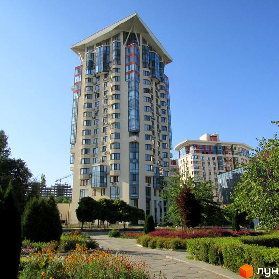 Житловий комплекс Сонячна брама: висока будівля світлого кольору з великими вікнами та похилим дахом. Перед будинком доглянутий парк з доріжками та квітами.