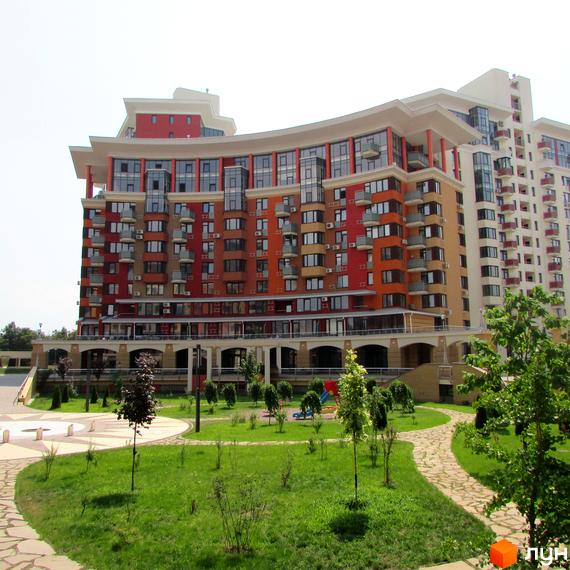 Житловий комплекс Сонячна брама з фасадом у відтінках червоного та помаранчевого, з численними вікнами та балконами. Перед будинком зелений газон, доріжки та молоді дерева.