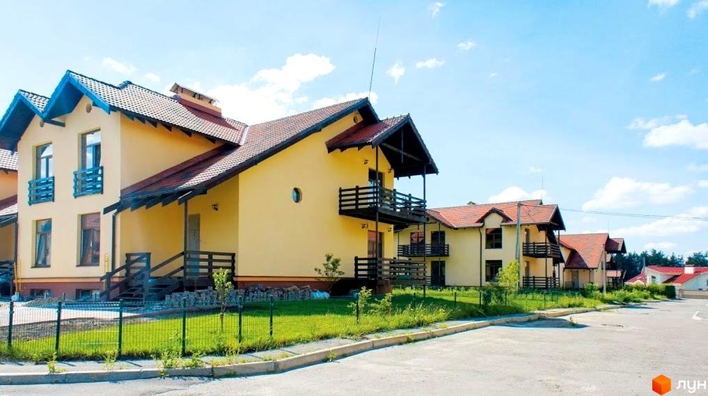 Зовнішній вигляд будинків житлового комплексу КМ Садок Вишневий з жовтою фасадною обробкою. Прибудинкова територія з зеленими насадженнями та паркувальними місцями.