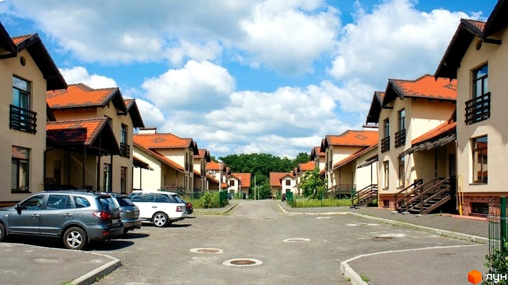 Зображення житлового комплексу з двоповерховими будинками з червоними дахами. На передньому плані асфальтована дорога з припаркованими автомобілями.
