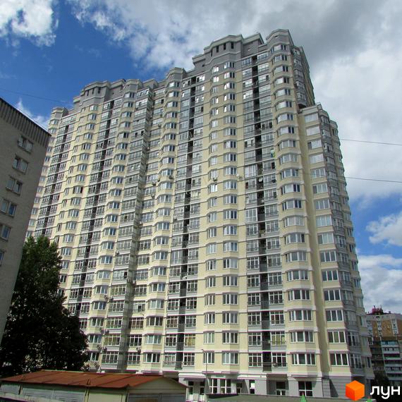 Багатоповерховий житловий будинок світлого кольору з численними вікнами та балконами. На передньому плані видно частину іншої будівлі та дерева.