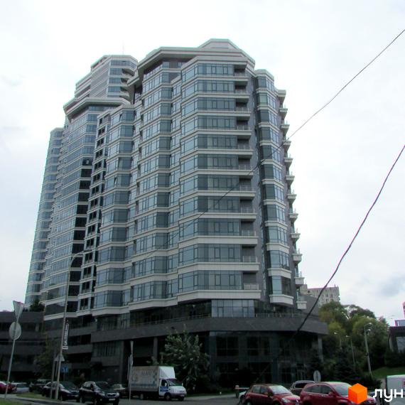 Високий житловий комплекс PecherSKY з багатокутними секціями, великими вікнами та балконами. Поруч проїжджа частина з автомобілями та зелені насадження.