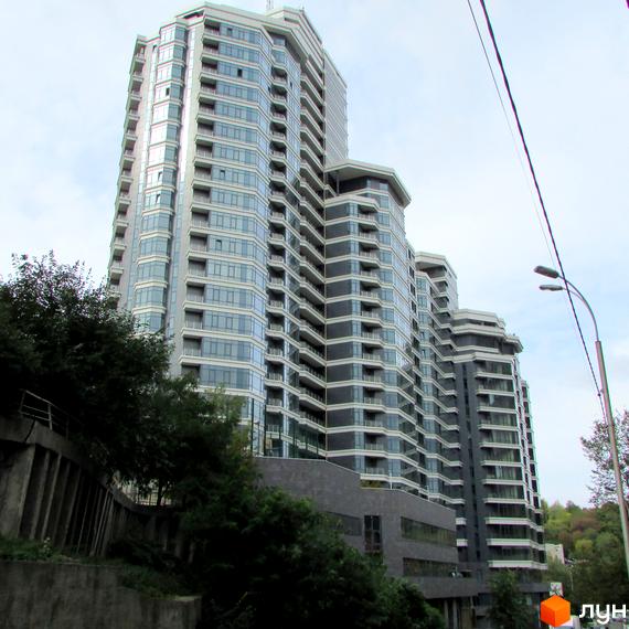 Житловий комплекс PecherSKY з багатосекційним фасадом, що поєднує скляні вікна та темні панелі. Навколо видно зелені насадження та бетонну стіну.