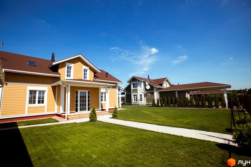 Будинок з жовтими стінами та коричневим дахом на тлі блакитного неба. Перед будинком доглянутий газон та доріжка.