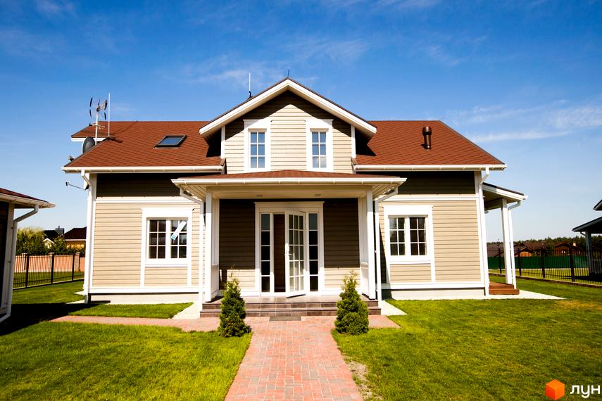 Зовнішній вигляд будинку в Котеджному містечку Бобриця-Парк. Будинок з коричневою черепицею та світлими стінами, перед будинком зелена галявина та доріжка з бруківки.