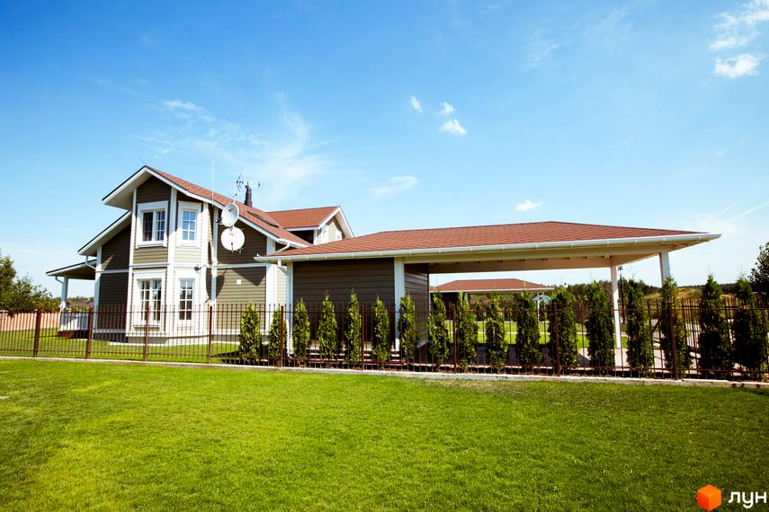 Двоповерховий будинок з коричневою черепицею та великими вікнами. На території зелена галявина, котеджний комплекс огороджений металевим парканом.