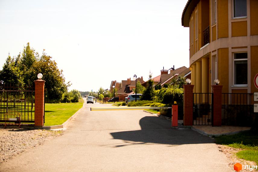 В'їзд до Містечко котеджів КМ Бобриця-Парк. На передньому плані шлагбаум та цегляні стовпи з ліхтарями. Далі видно будинки з черепичними дахами та доглянутою прибудинковою територією.