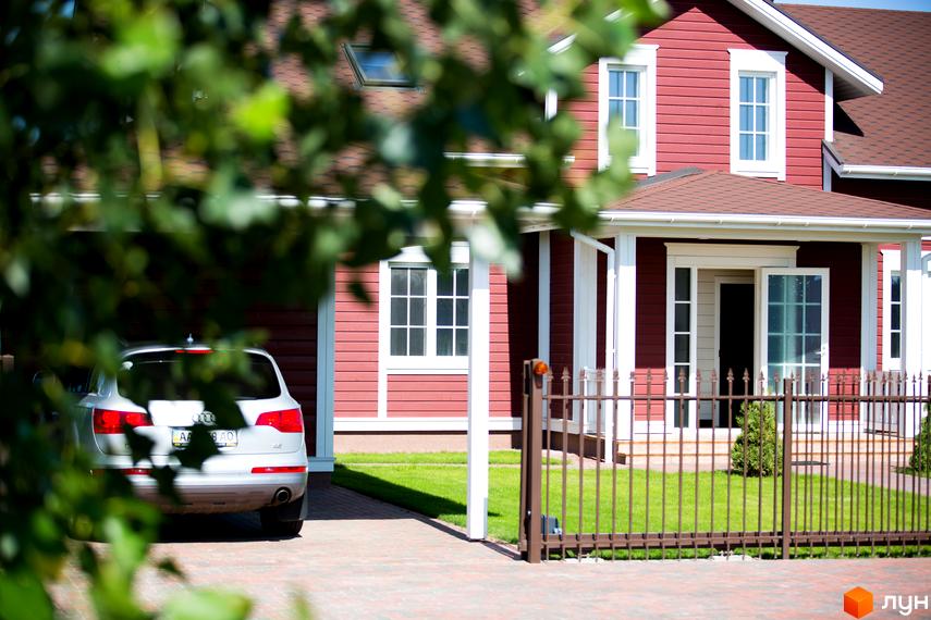 Будинок з червоним фасадом та білими віконними рамами. Перед будинком доглянута прибудинкова територія з газоном та кованим парканом. Містечко котеджів Бобриця-Парк.