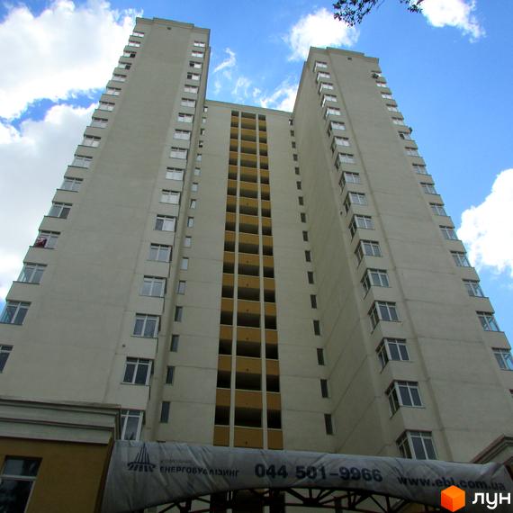 Високий житловий комплекс ЖК Затишний дім з багатьма вікнами та балконами на фоні блакитного неба з хмарами. На фасаді видно частину будівельного проєкту та номер телефону.