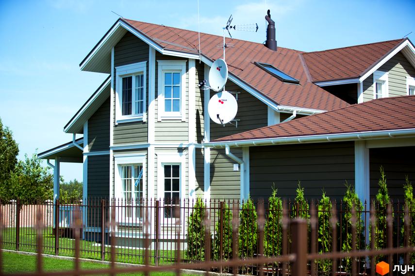 Двоповерховий будинок з коричневою черепицею та світлими вікнами. Перед будинком розташований кований паркан та зелена трава.