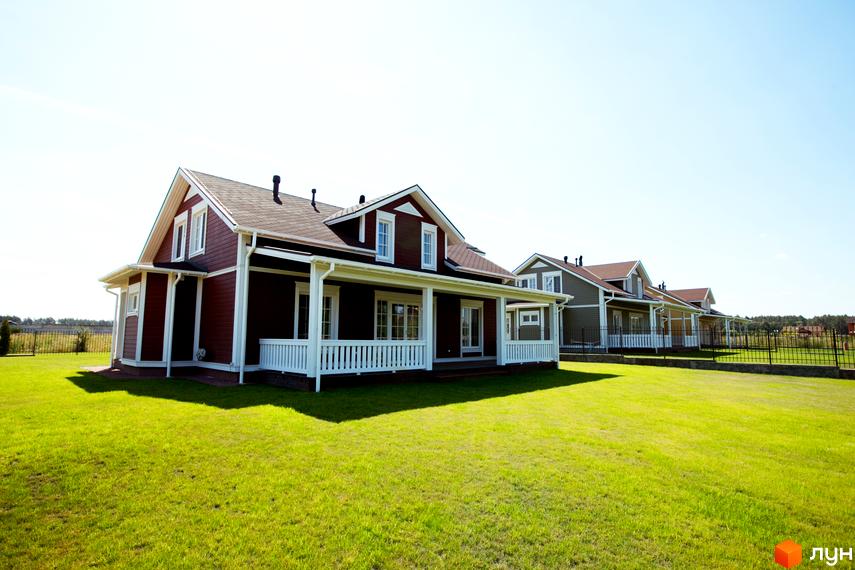 Будинок з темно-бордовими стінами та білими колонами на зеленому газоні. Поруч розташовані інші будинки в котеджному містечку КМ Бобриця-Парк.
