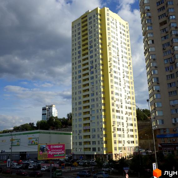 Житловий комплекс на Саперно-Слобідській, 24 - висотна жовто-біла новобудова з численними балконами. Навколо розташовані інші будинки та парковка.