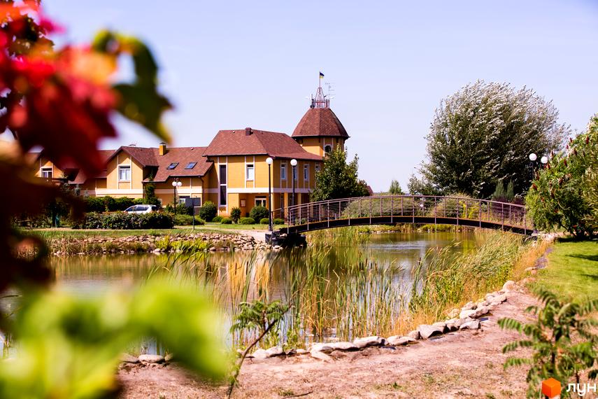 Житловий комплекс КМ Бобриця-Парк з жовтими стінами та коричневим дахом. На території є місток через водойму та зелені насадження.