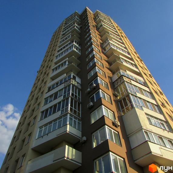 Вид на висотний житловий комплекс на вул. Ушинського, 14б. Будинок має коричнево-бежевий фасад, численні балкони та великі вікна. Навколо будинку видно блакитне небо та невеликі хмари.