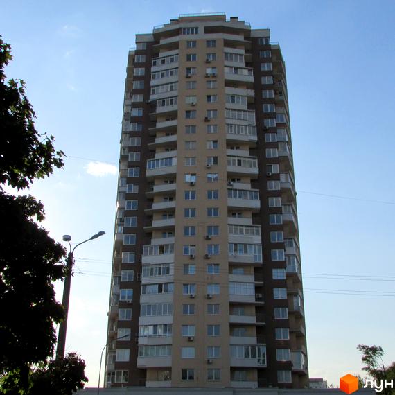 Висотна новобудова на вулиці Ушинського, 14б з численними балконами та вікнами. Будинок має світло-бежевий та темно-коричневий фасади. Навколо видно дерева та стовп з ліхтарем.