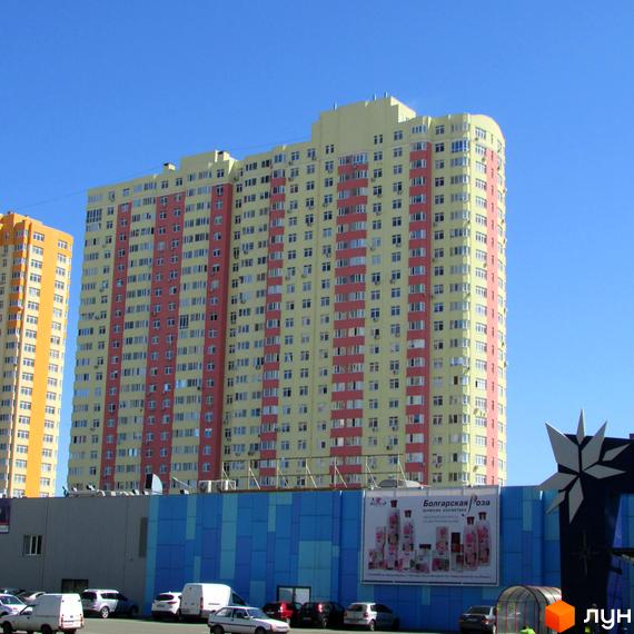 Житловий комплекс Яскравий у жовто-рожевих кольорах з численними вікнами та кондиціонерами. На передньому плані видно парковку та частину фасаду будівлі з синіми панелями.