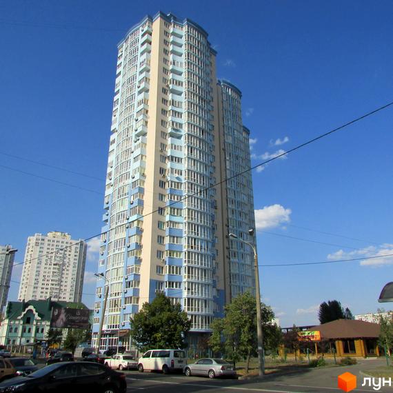 Житловий комплекс Озерні зорі – висотна новобудова з блакитними та бежевими фасадами, розташована на фоні блакитного неба. Поруч видно дерева та дорогу з автомобілями.