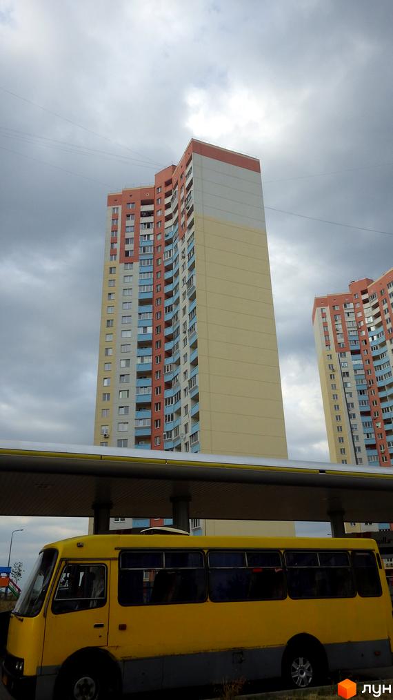 Житловий комплекс на вул. Милославська, 2, 2б, 2в. Багатоповерхові будинки з різнокольоровими фасадами та балконами на тлі хмарного неба. Перед будинками видно жовтий автобус під навісом.