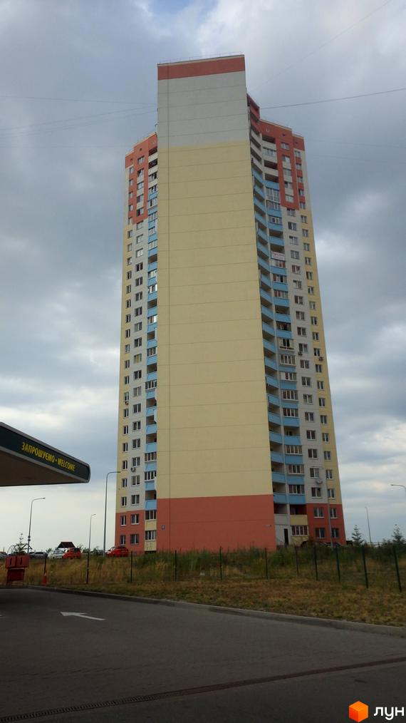 Багатоповерховий житловий комплекс на вул. Милославська, 2, 2б, 2в. Будинок має секції різних кольорів: бежевий, червоний, білий. Навколо будинку видно дорогу та зелені насадження.