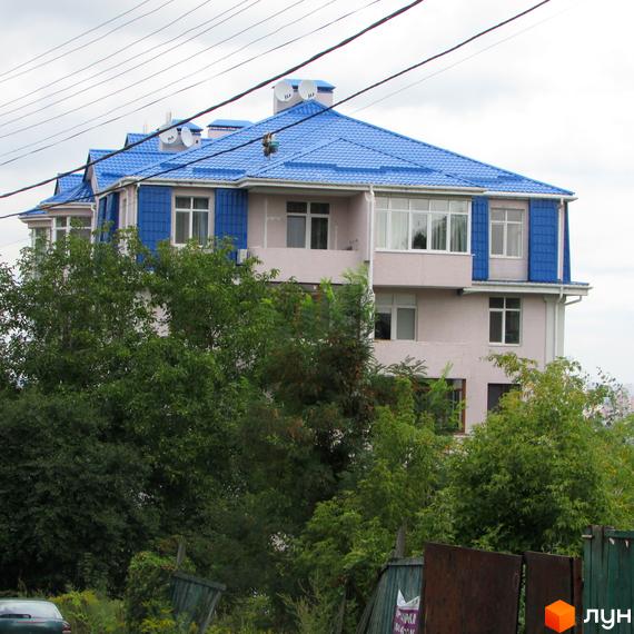 Зовнішній вигляд житлового комплексу пров. Іллі Рєпіна (Шишкінський) з блакитним дахом та світло-рожевими стінами. Навколо будинку зелені дерева та огорожа.