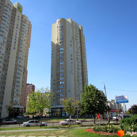 Високі житлові будинки на вулиці Драгоманова (будмайданчики 1, 2, 3) під блакитним небом. Перед будинками розташована дорога з автомобілями та зелена зона з квітами.