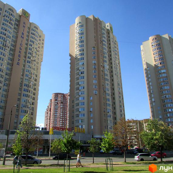 Високі світло-бежеві багатоповерхові будинки з численними вікнами та кондиціонерами. Перед будинками зелена зона з деревами та доглянутим газоном, дорогою та припаркованими автомобілями.