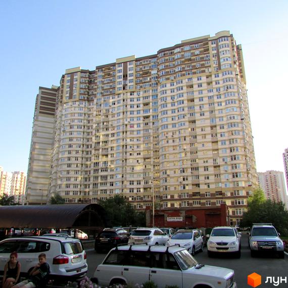 Новобудова на Ахматової, 22: високий будинок світлих відтінків з коричневими верхніми поверхами. Біля нього розташований паркінг з автомобілями.