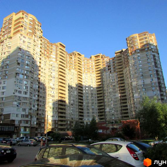 Багатоповерховий житловий будинок з хвилястим фасадом та світло-коричневими секціями. Перед будинком розташований паркінг з автомобілями.