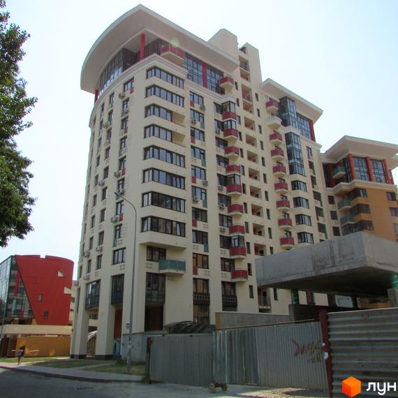 Житловий комплекс Сонячна брама: високий будинок світлого кольору з численними вікнами та червоними балконами. На передньому плані дорога та паркан.