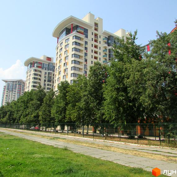 Багатоповерхові будинки Житлового комплексу Сонячна брама світлого кольору з вигнутими дахами. Перед ними зелені дерева, доріжка та газон.