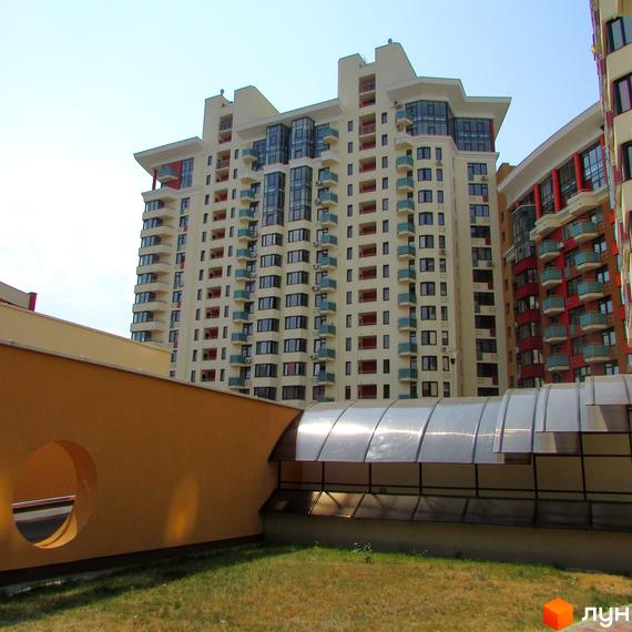 Житловий комплекс Сонячна брама: багатоповерховий будинок світлого кольору з балконами. Прилегла територія з газоном та прозорим накриттям.