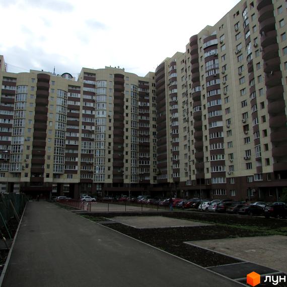 Житловий комплекс Парковий з бежевими стінами та коричневими балконами, що утворює вигнуту форму. На передньому плані доріжка, зелені насадження та парковка з автомобілями.