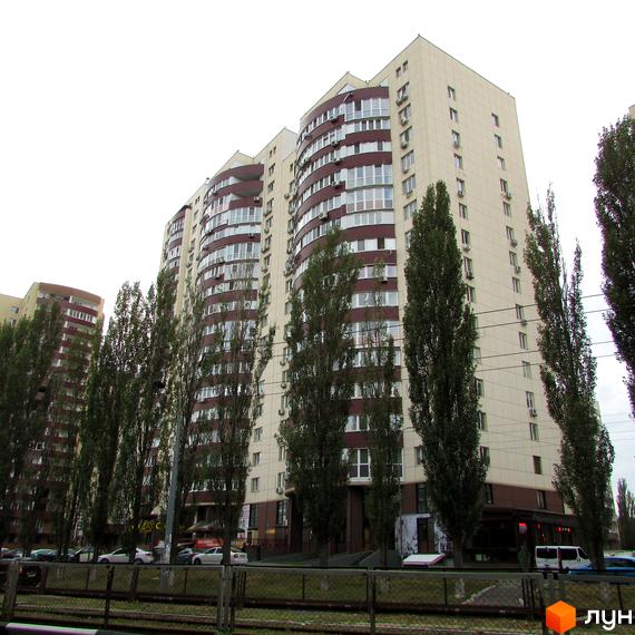 Житловий комплекс Парковий: багатоповерховий будинок світлого кольору з темними акцентами та великими вікнами. Перед будинком ростуть високі дерева та проїжджа частина.