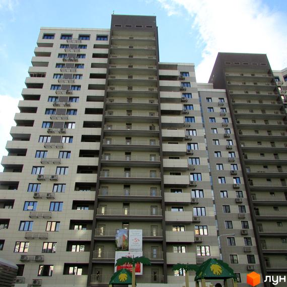 Зовнішній вигляд багатоповерхового житлового комплексу ЖК Островського, 40. Фасад будівлі оздоблений світлими панелями та темними вставками, з численними балконами та вікнами. На передньому плані видно елементи дитячого майданчика.