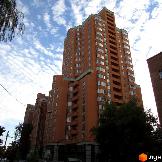 Житловий комплекс Златоуст – це цегляна новобудова з вигнутими фасадами та численними балконами, що має багаторівневу архітектуру. Навколо будівлі ростуть дерева, а на задньому плані видно хмарне небо.