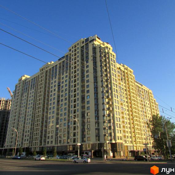 Житловий комплекс Венеція - висотна жовта новобудова з численними балконами та вікнами на фоні блакитного неба. Біля будинку розташована дорога з автомобілями та пішоходами.