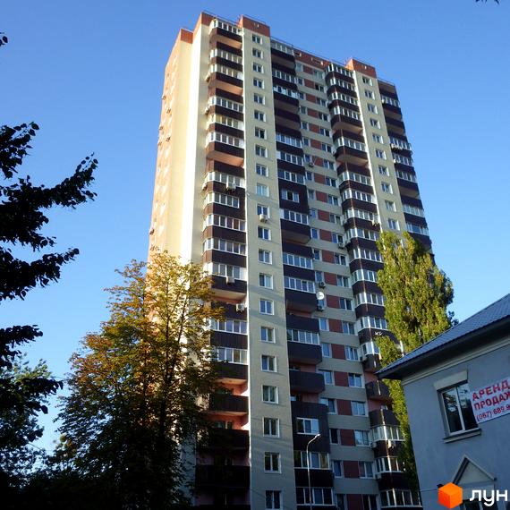 Висотна новобудова Будинок на Шулявці з балконами та кондиціонерами на фоні блакитного неба. Поруч дерева та частина будівлі з написом.