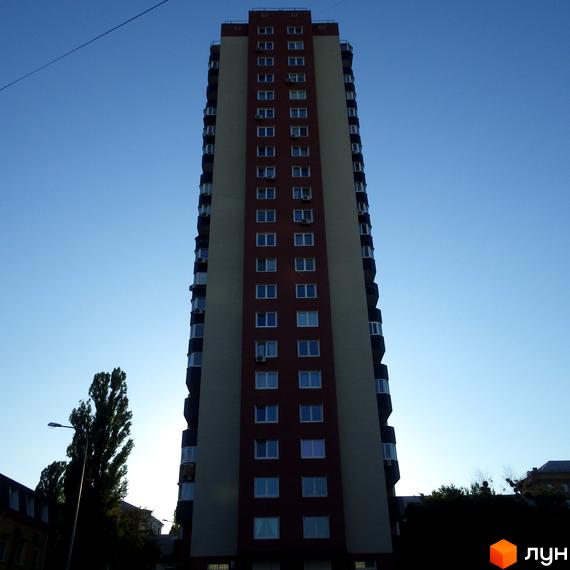 Високий житловий комплекс Будинок на Шулявці з червоно-бежевим фасадом та численними вікнами. Навколо видно дерева та блакитне небо.