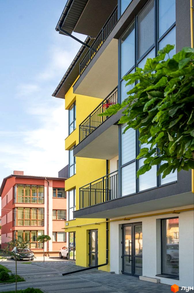 Фасад житлового комплексу Італійський з жовтими та рожевими стінами, балконами та великими вікнами. Поруч розташована прибудинкова територія з парковкою та зеленими насадженнями.